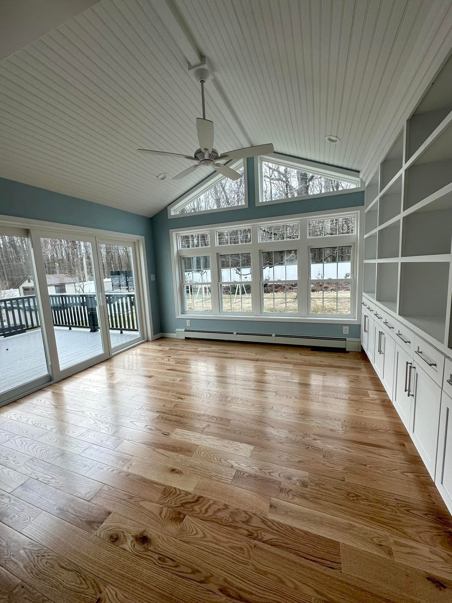 Bright sunroom with hardwood floors, large windows, and vaulted white ceiling overlooking a deck and trees