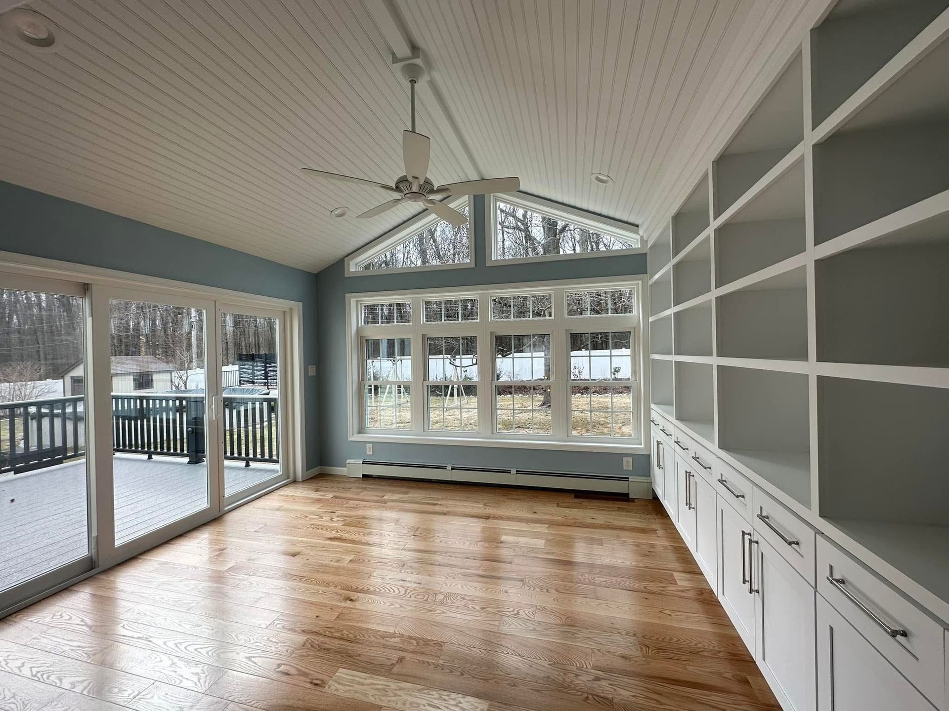 Bright sunroom with hardwood floors, white built-in cabinets, large windows, and a ceiling fan.