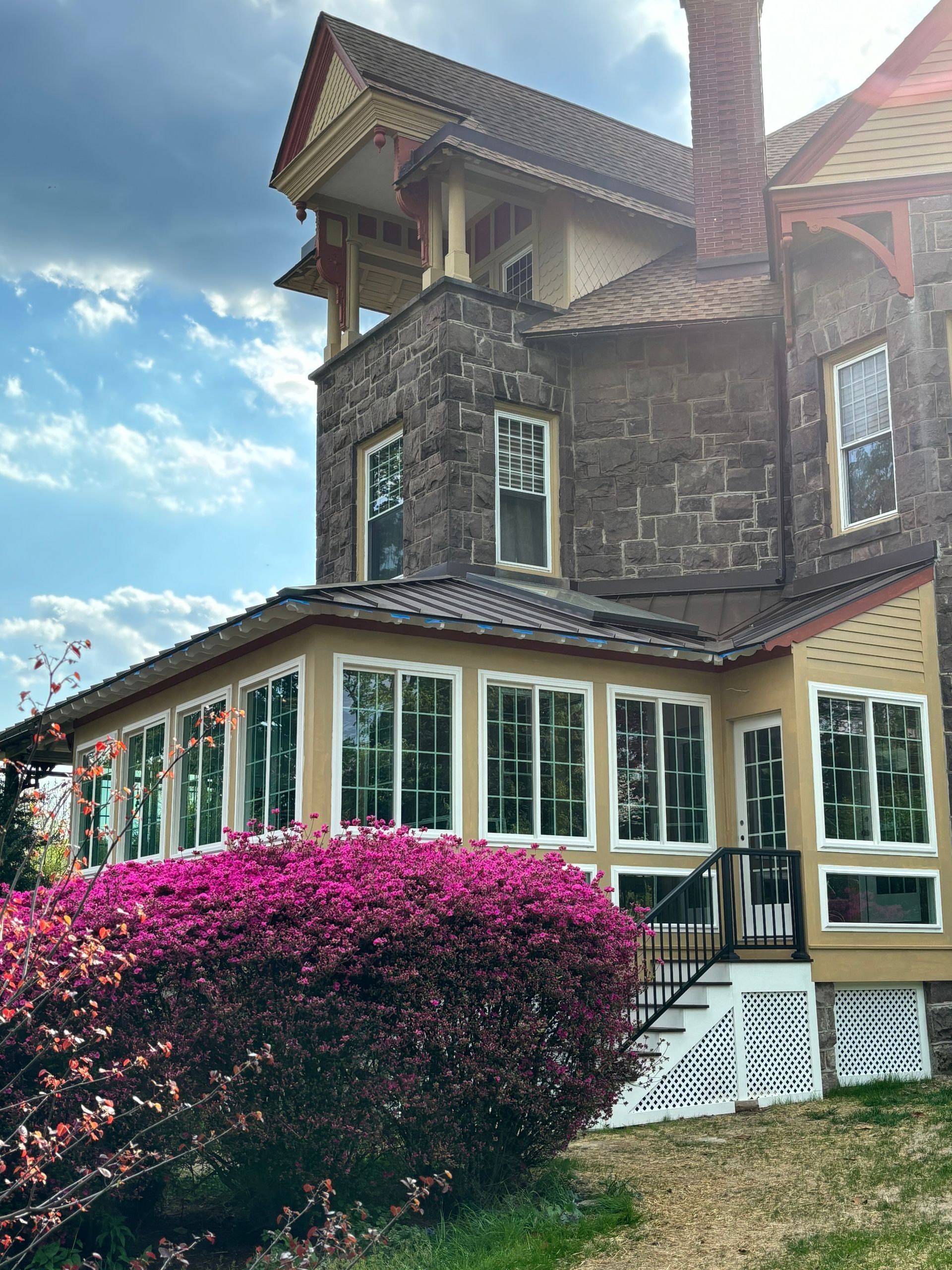 Stone house with white-trimmed windows, red roof, and pink flowering bush in front under a blue sky