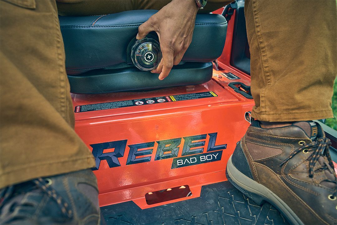 Person adjusting the seat on a red Bad Boy Rebel lawnmower, showing its control panel.