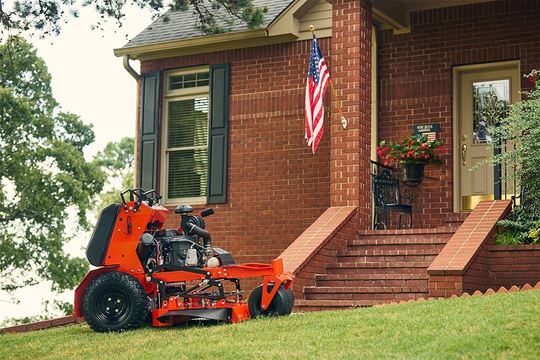 Orange zero-turn mower on a lawn in front of a brick house with an American flag.