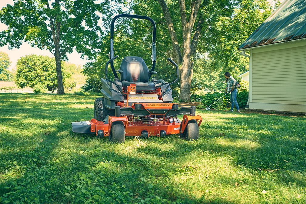 Orange and black zero-turn mower on green grass. A person is tending to bushes near a light green building.