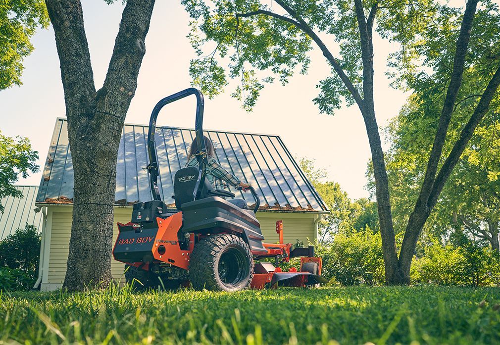 A person on a red and black zero-turn mower cuts grass near a house and trees.