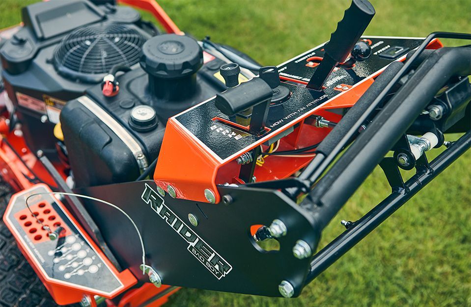 Close-up of a zero-turn lawnmower's control panel. Black, orange, and gray, with levers, pedals, and gauges. The word 