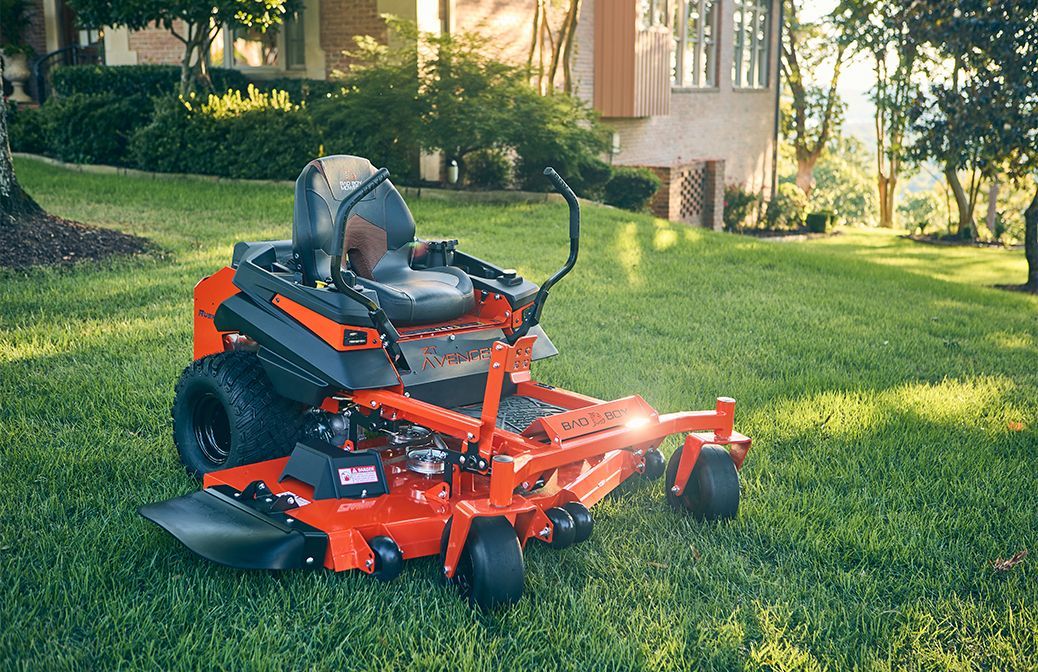 Orange zero-turn lawn mower on green grass. House and trees in the background. Sunny outdoor setting.