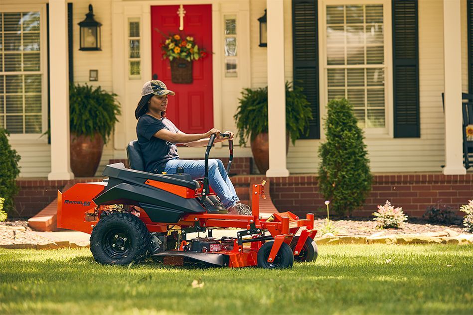 Woman on orange zero-turn mower cutting grass in front of white house with red door.
