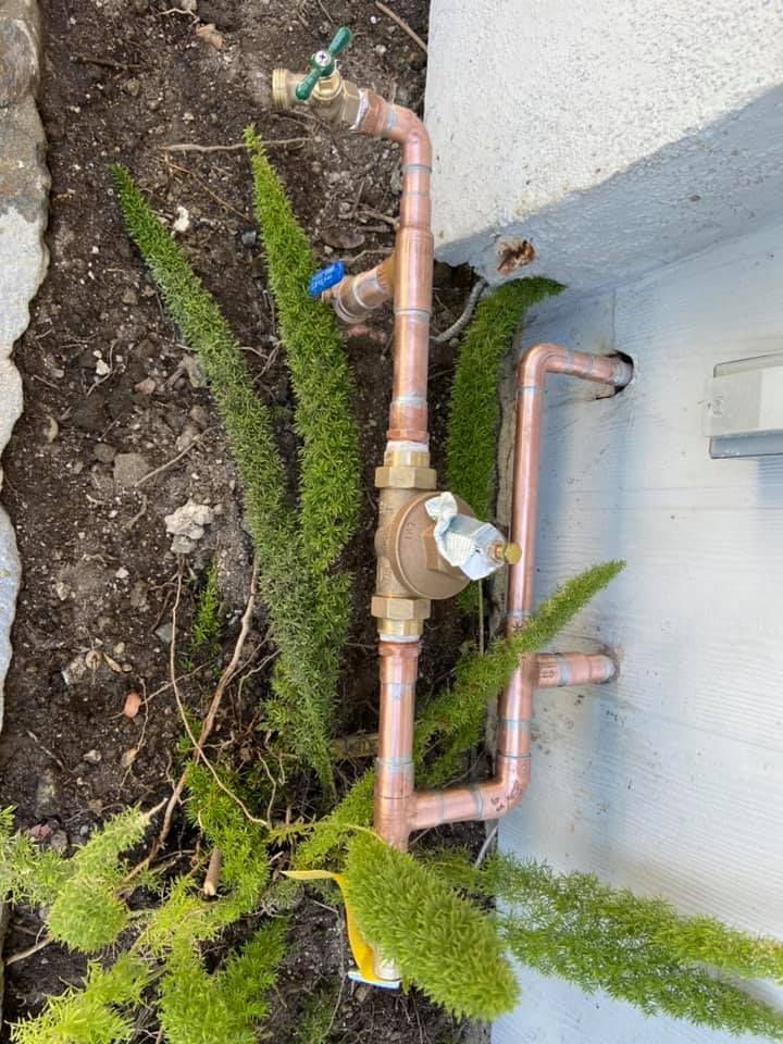 Copper piping with a pressure regulator and two spigots installed against an exterior wall near soil and green plants.