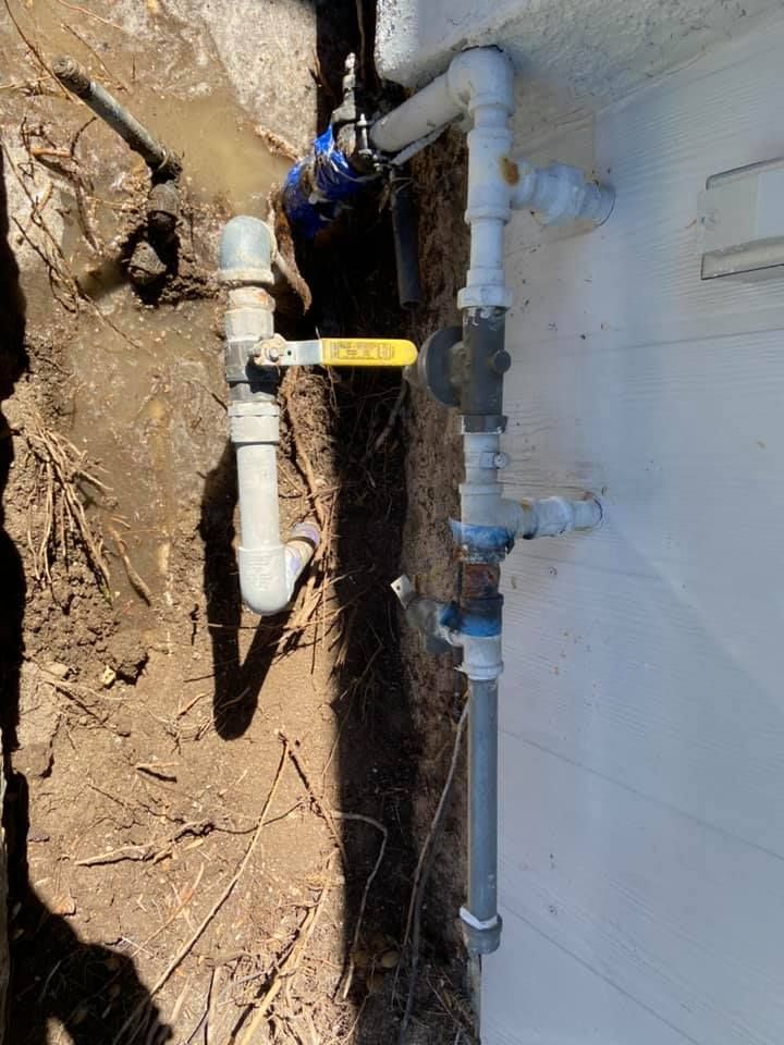 Outdoor plumbing pipes in a dirt trench beside a house wall, with a yellow valve handle.