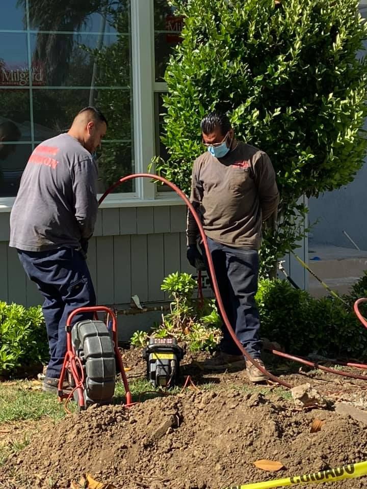 Two workers use a hose and power tool to landscape a dirt bed beside a house.
