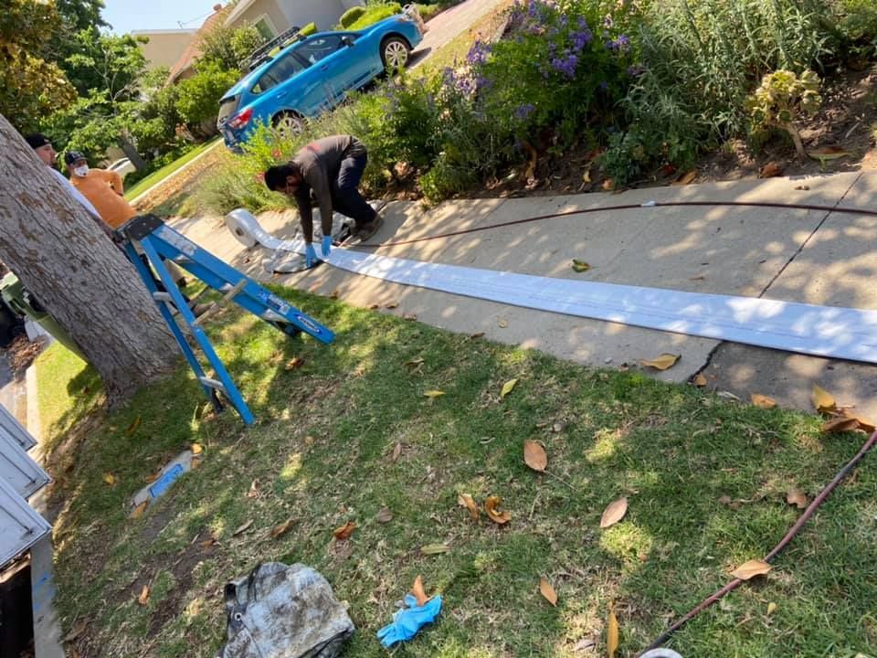Person kneels on a sidewalk beside a long white strip, with a blue ladder and truck in a sloped yard.