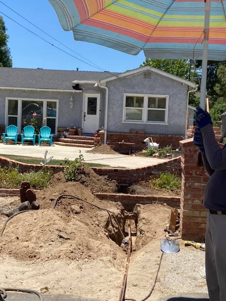 House under construction with dug-up yard, patio chairs, and a person holding a striped umbrella
