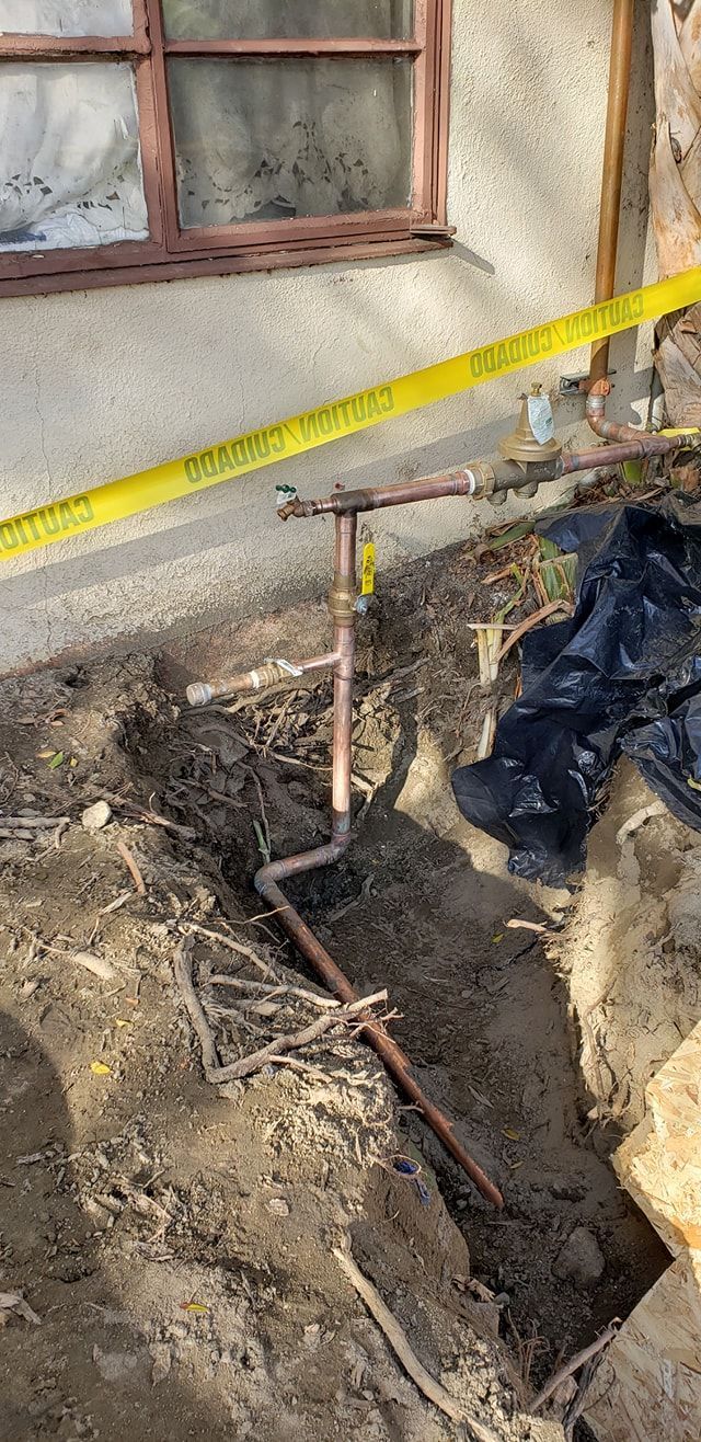 Backyard excavation beside a house, with exposed pipes, dirt trench, and caution tape
