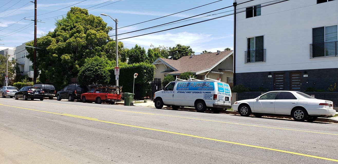 Parked cars and a delivery van along a sunny residential street beside a white building and trees