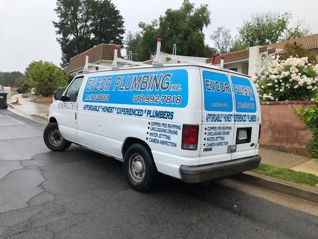 White service van with blue lettering parked on a wet suburban street by a brick wall and trees.