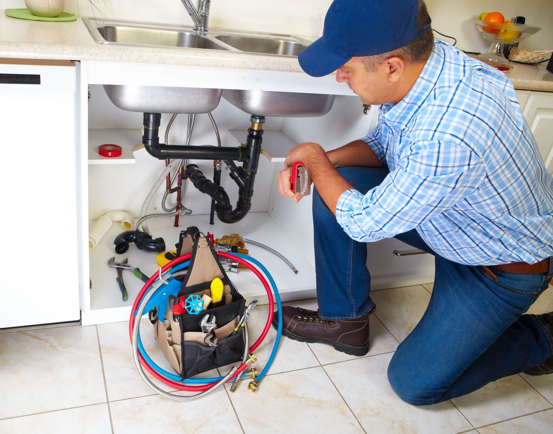 Plumber kneeling under a sink, inspecting pipes with tools and coiled colored hoses on the floor.