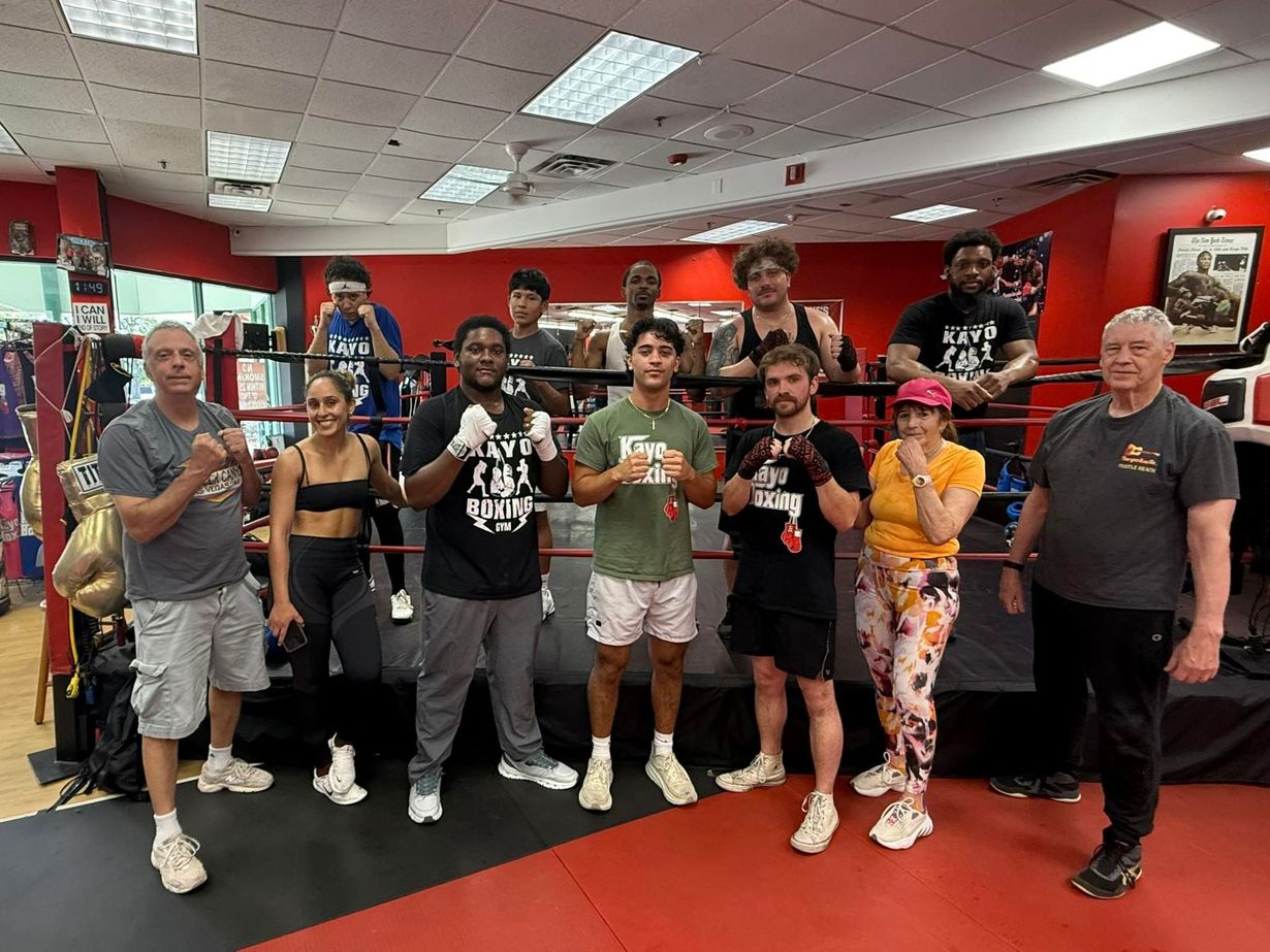 People posing inside a boxing ring. Some are wearing boxing gloves. Red and black ring. Gym setting.