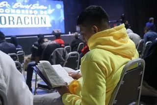 A man in a yellow hoodie is sitting in a chair reading a bible.