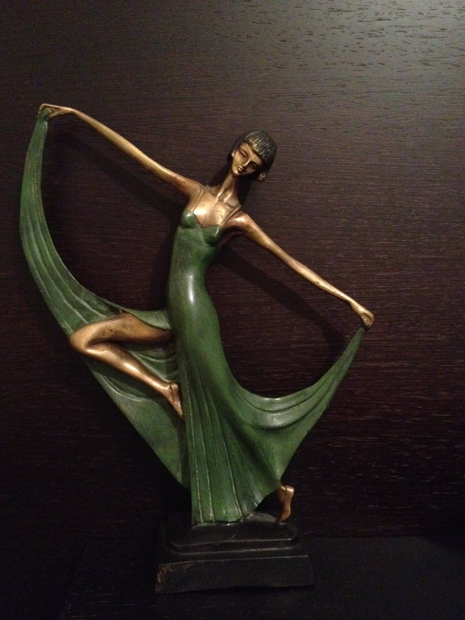 Art Deco-style bronze statue of a dancing woman in green dress with arms outstretched, on a dark background.