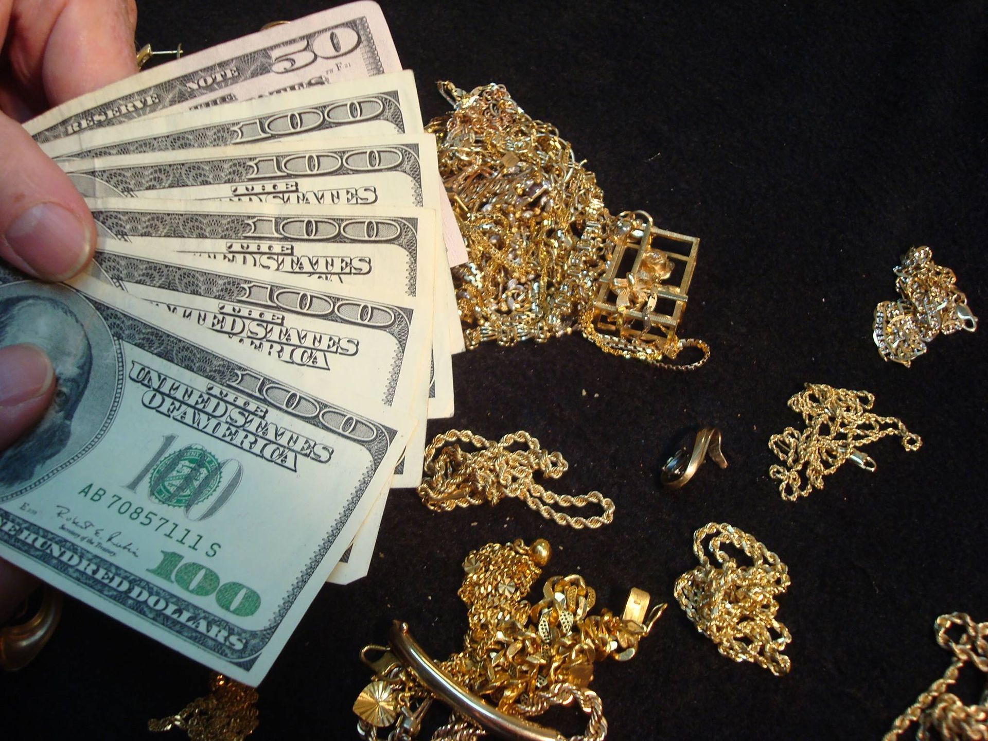 Hand holding cash next to scattered gold jewelry on a dark surface.