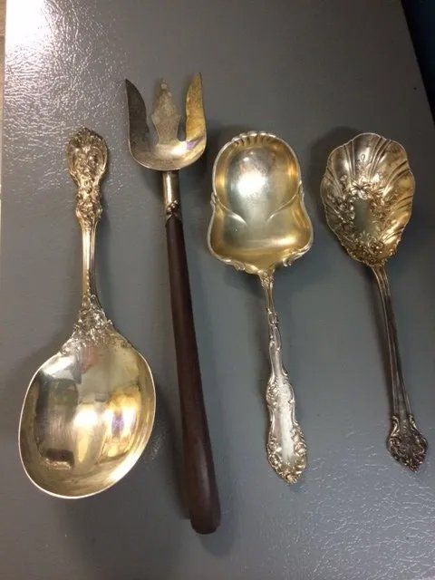 Four antique silver serving utensils on a grey surface; a large spoon, serving fork, ladle, and small serving spoon.
