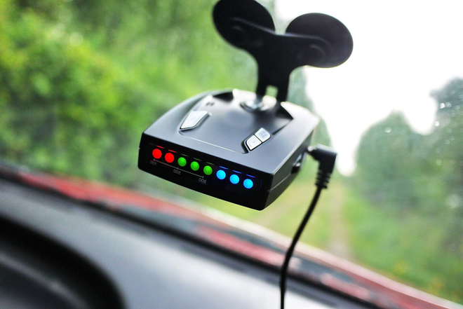 Small dashboard device with colorful LED lights mounted on a car windshield against a blurred green road backdrop