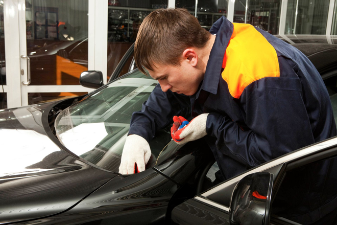 Mechanic inspecting a black car hood in a garage, wearing gloves and a navy jacket with orange shoulders