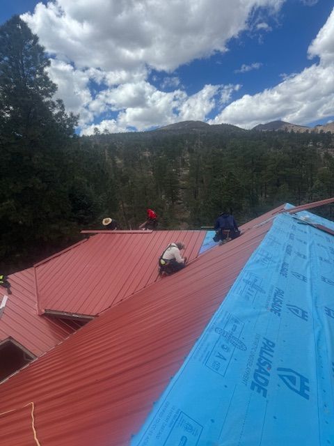 Workers on a red metal roof with blue underlayment, surrounded by pine trees and mountains under a cloudy sky