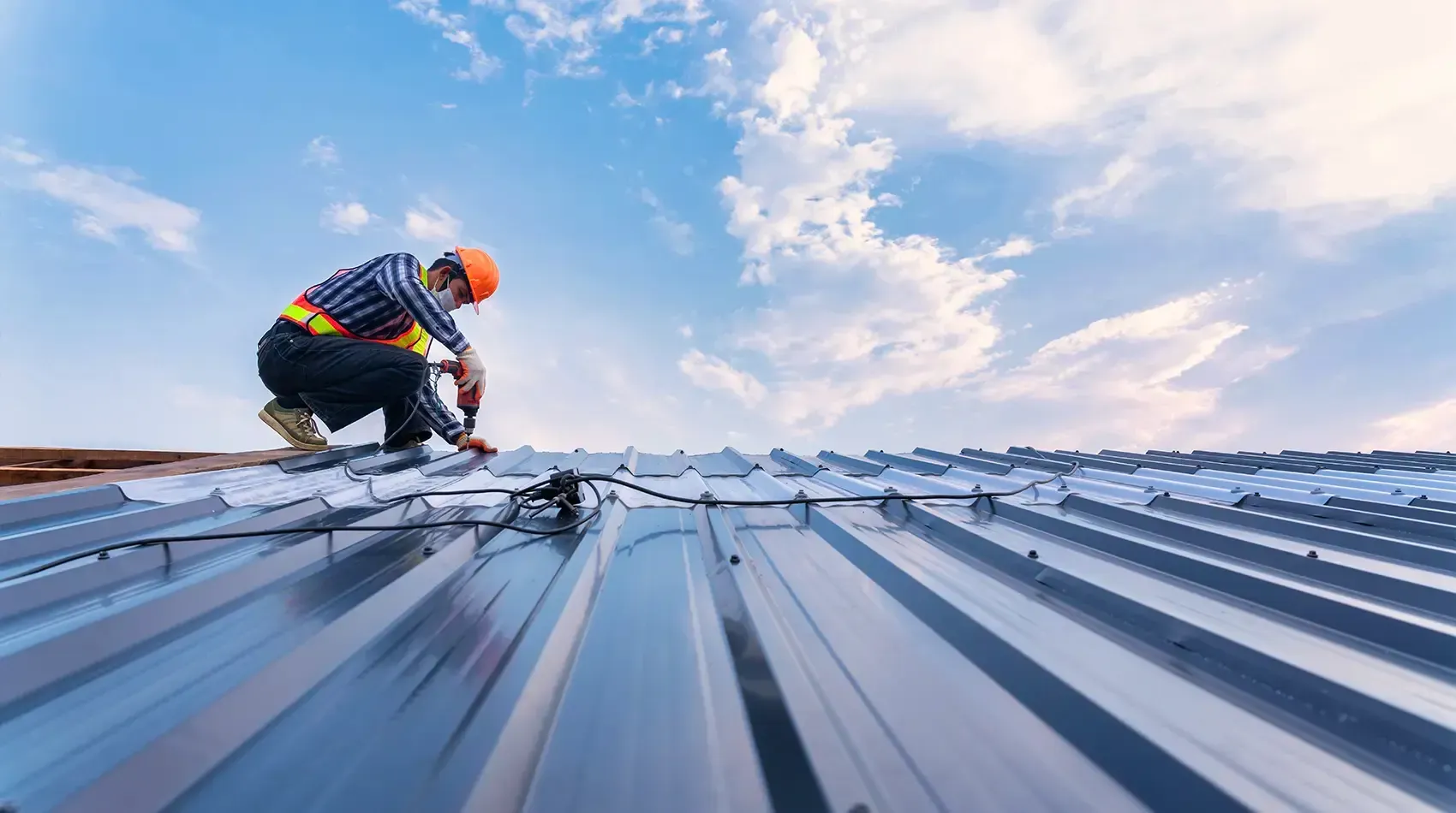 Worker on a metal roof against a blue sky, installing roofing panels with safety gear
