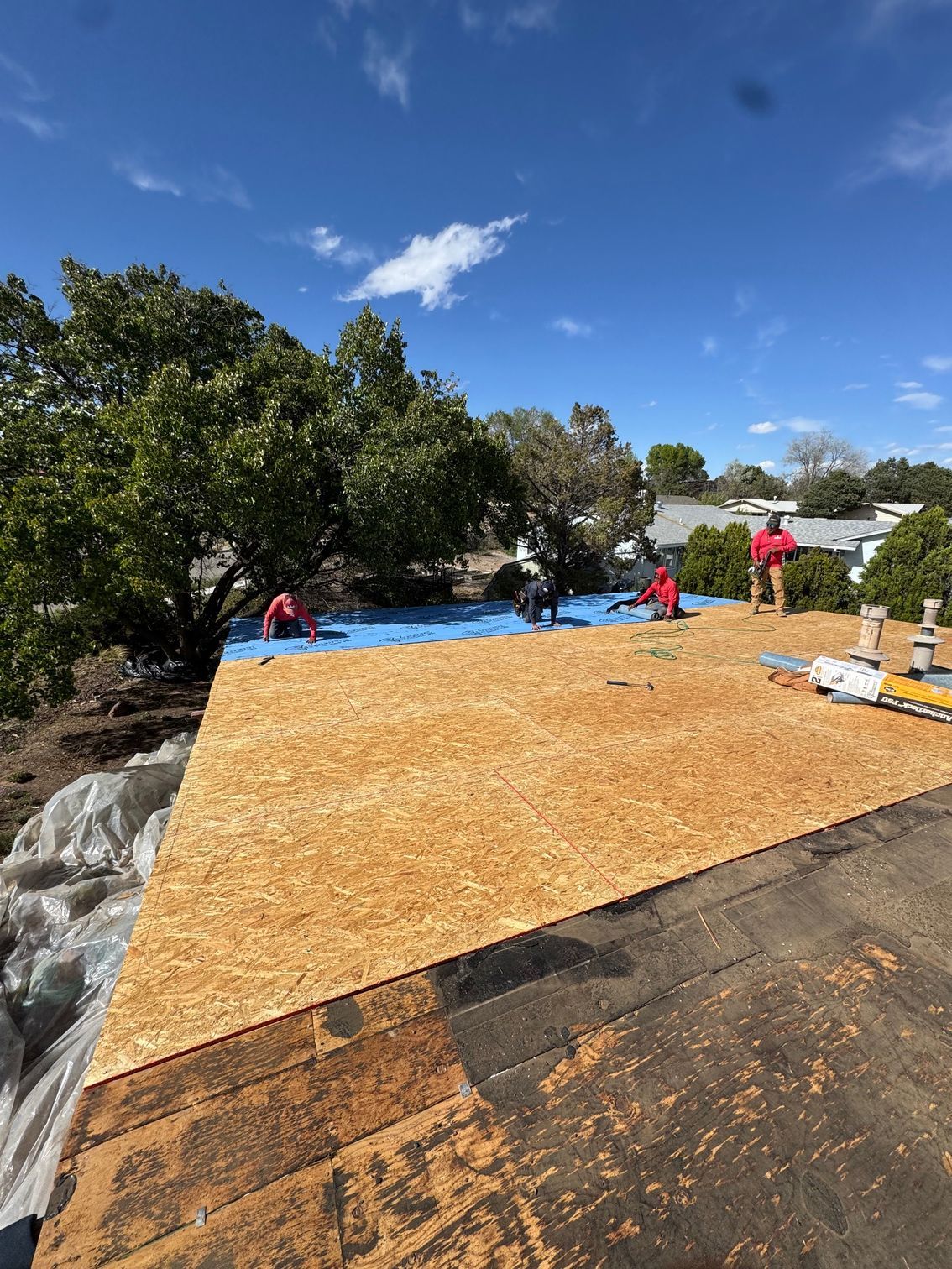 Workers installing plywood on a rooftop under a blue sky, with trees and a red jacket nearby.