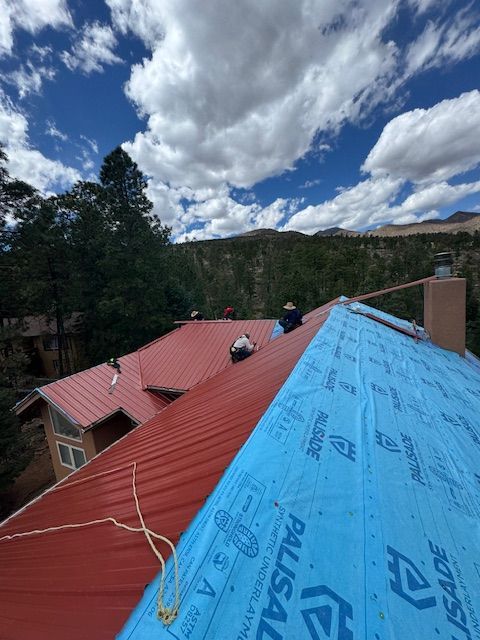 Roof under repair with blue and red roofing sheets beside a wooded hillside under a cloudy sky