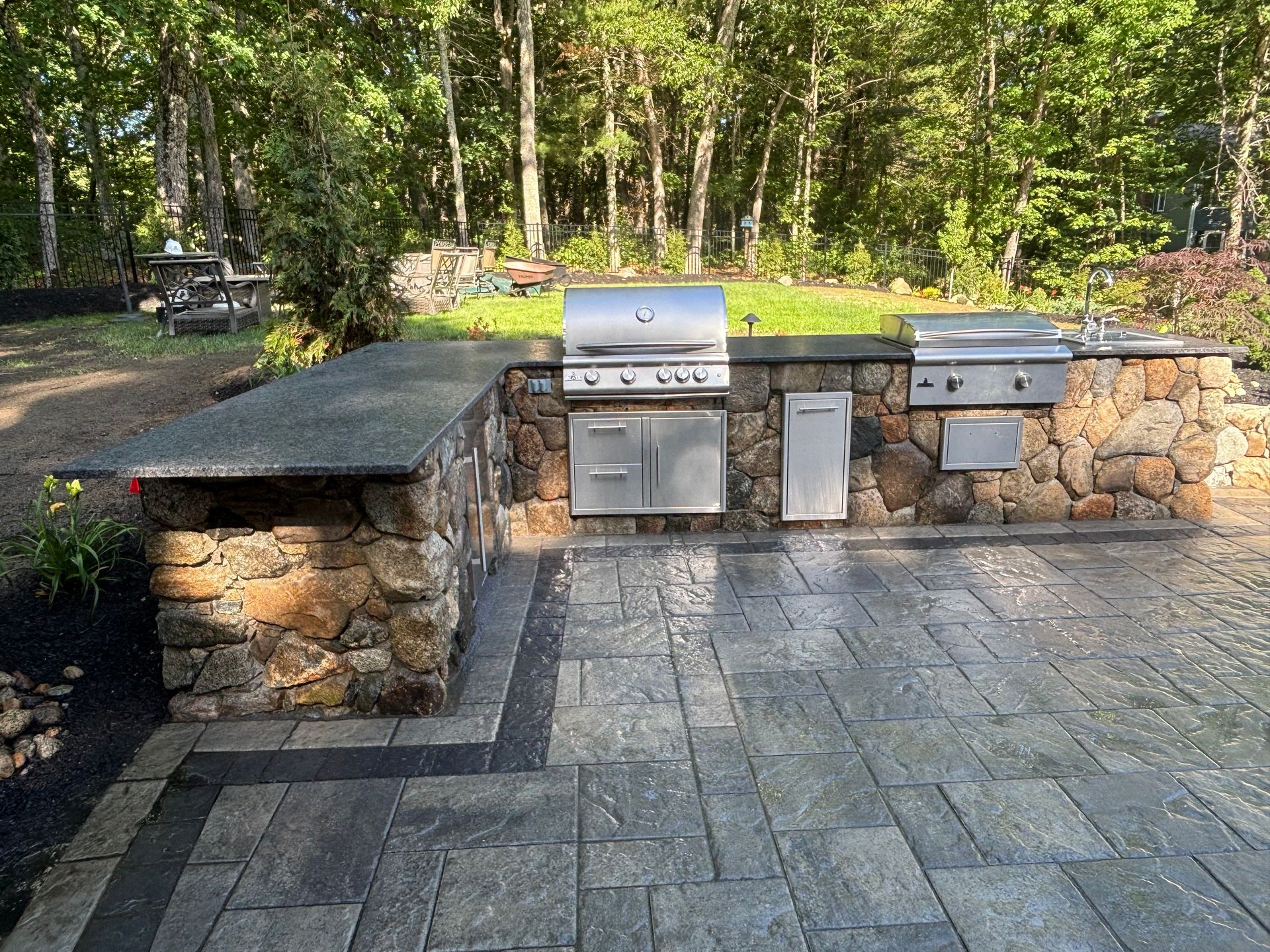 Outdoor stone patio with built-in stainless steel grill and stone counter seating in a wooded backyard