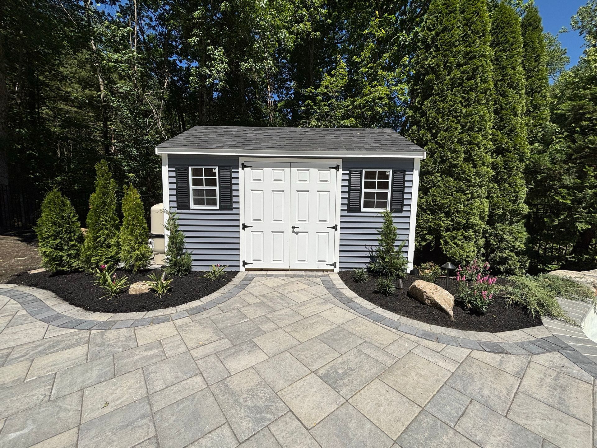 Small gray garden shed with white doors on a stone patio, surrounded by landscaping and trees.