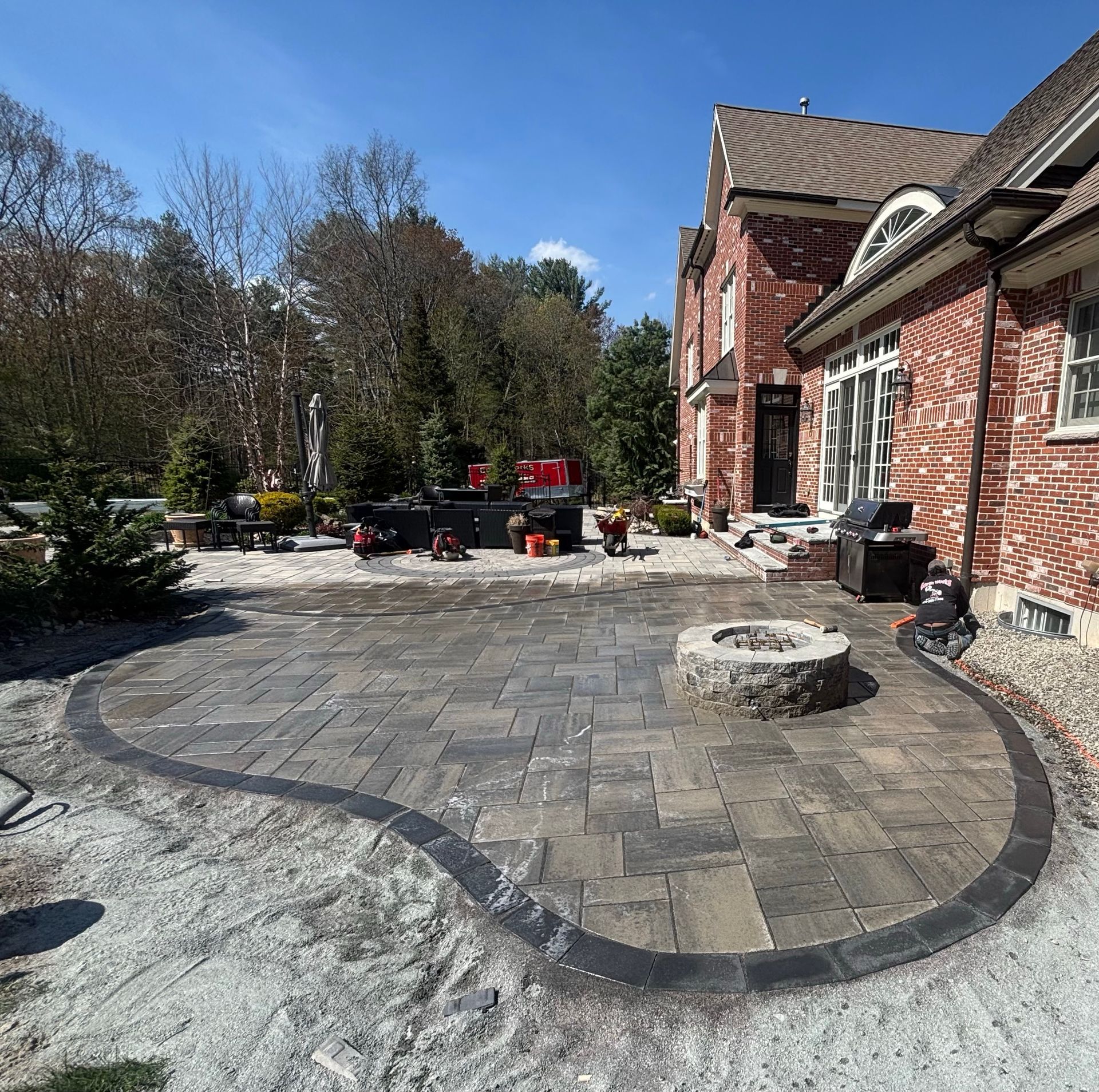 Brick patio with curved dark border beside a red brick house, patio furniture, and trees under a blue sky