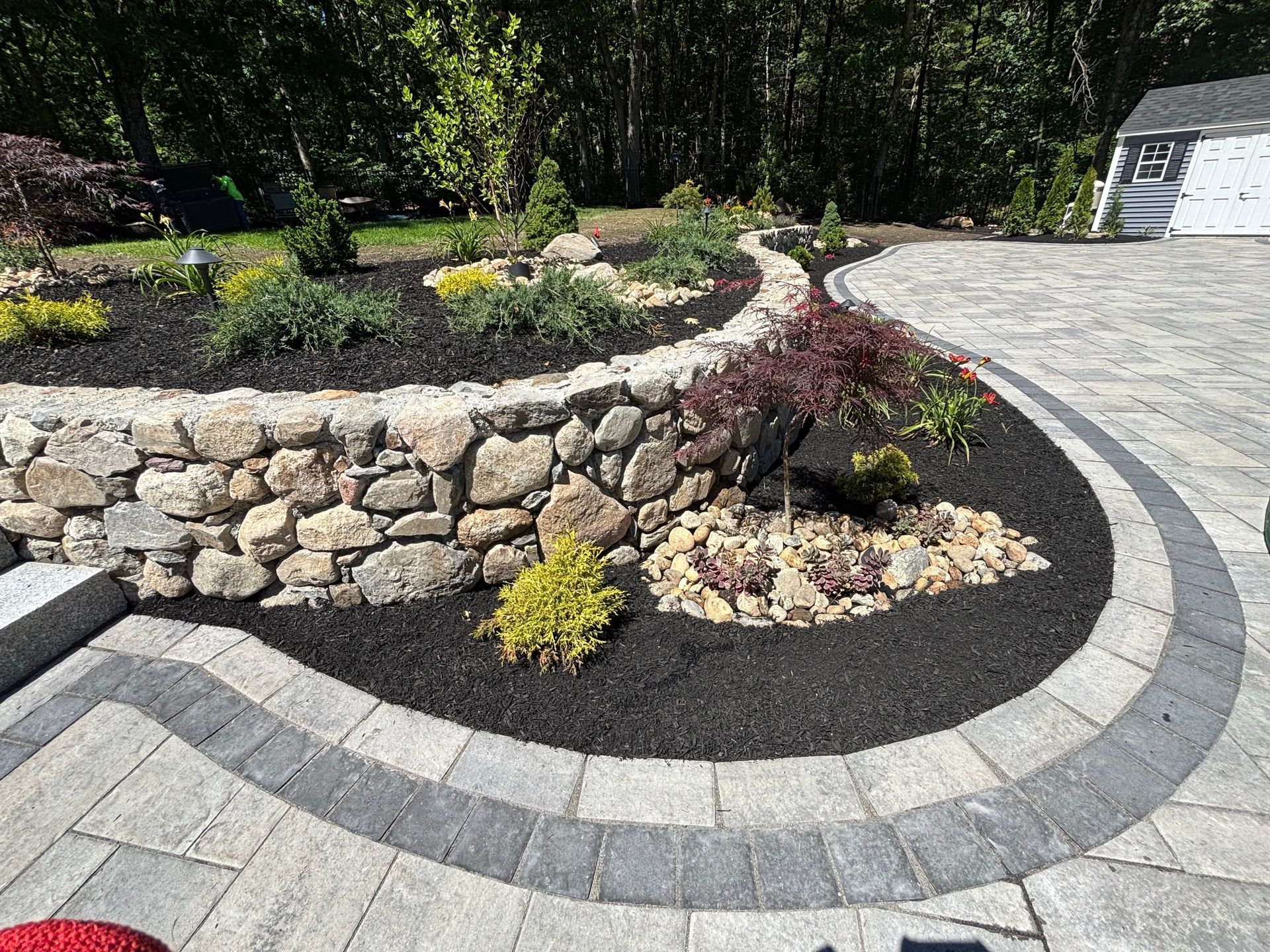 Curved stone retaining wall with mulch and shrubs beside a gray paver driveway and house driveway entrance