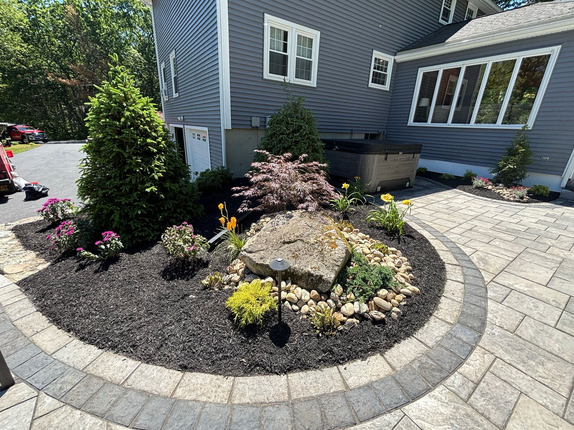 Curved stone-edged garden bed with rocks, flowers, and shrubs beside a gray house and paved patio