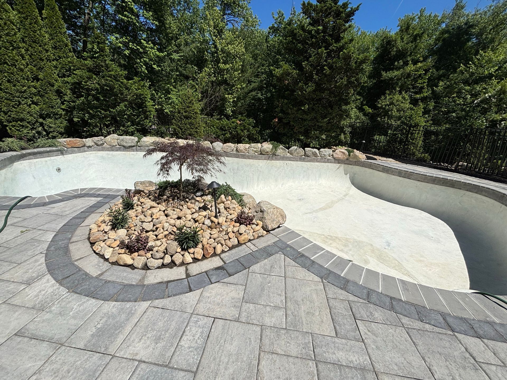Empty white concrete pool with a stone-lined planter and paved patio, backed by green trees.