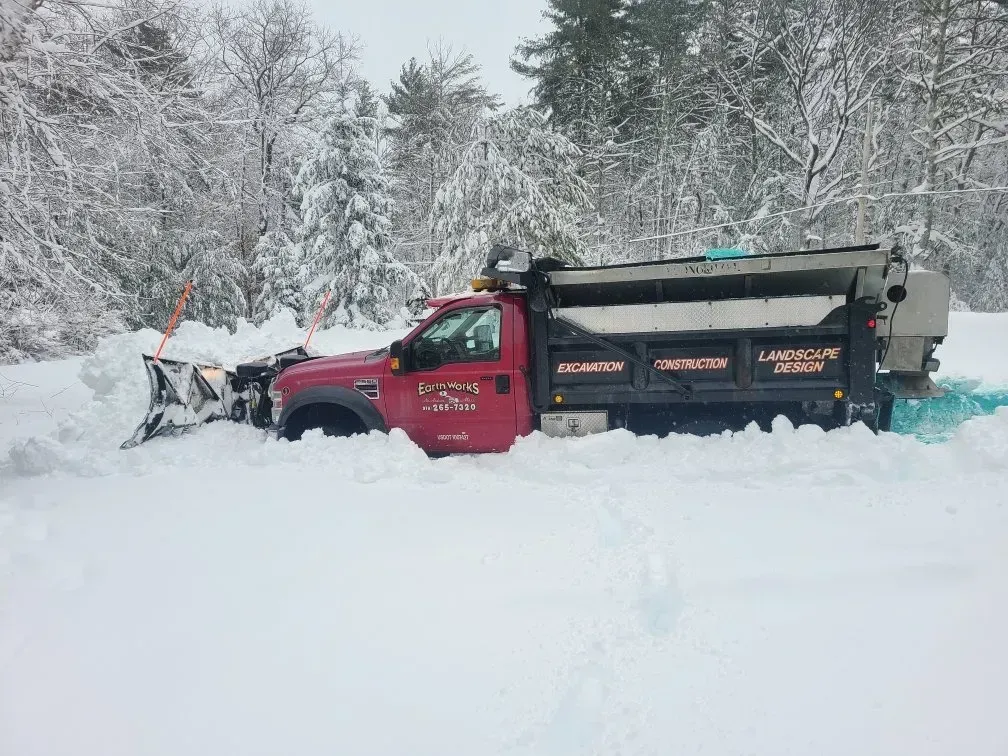 A red dump truck with a snow plow attached is parked in a deep, snow-covered landscape surrounded by evergreen trees.
