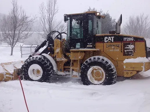 A yellow Caterpillar IT28G wheel loader with a plow attachment, parked in a snow-covered area during a light snowfall.