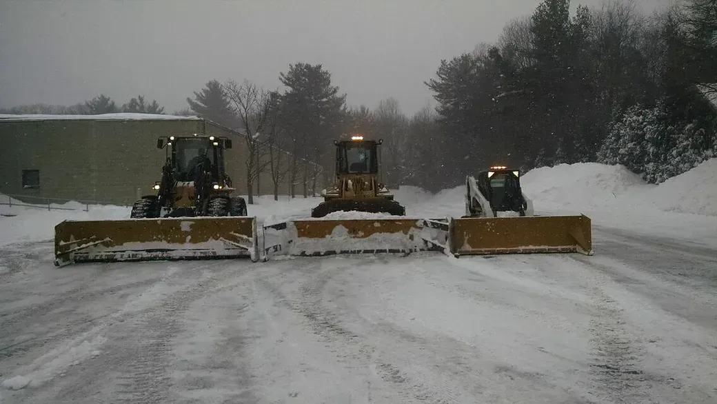 Three yellow snow-clearing vehicles lined up side-by-side on a snow-covered ground with a building and trees in the back.