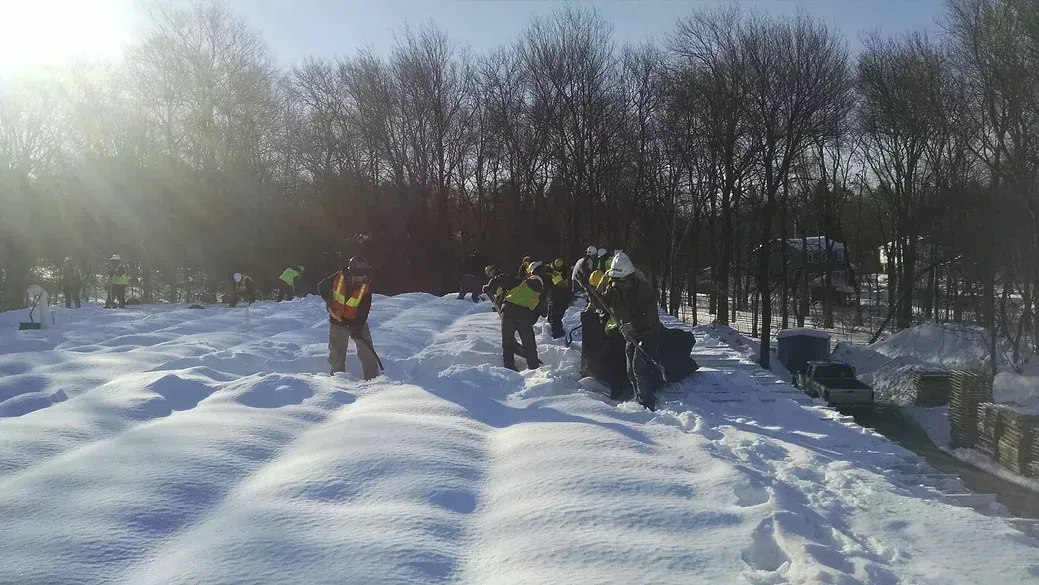 A group of workers in high-visibility vests walk through rows of snow in a park on a sunny day.