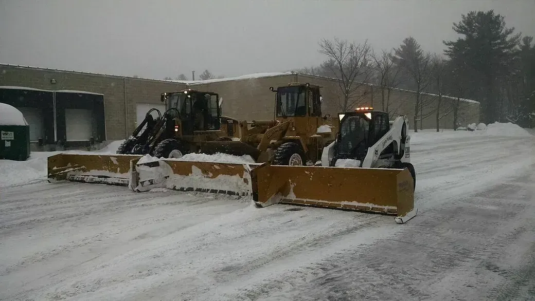Two yellow construction vehicles with snow plows attached to their fronts, parked in a snow-covered parking lot.