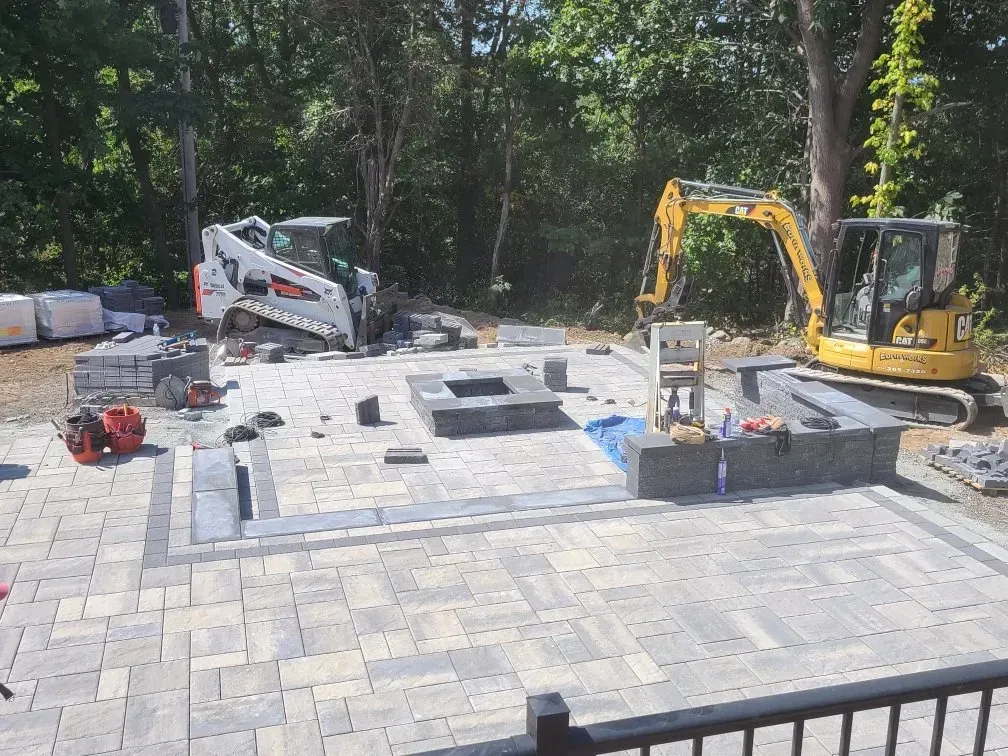 A white skid steer and yellow excavator work on a stone patio featuring a built-in fire pit in a backyard setting.