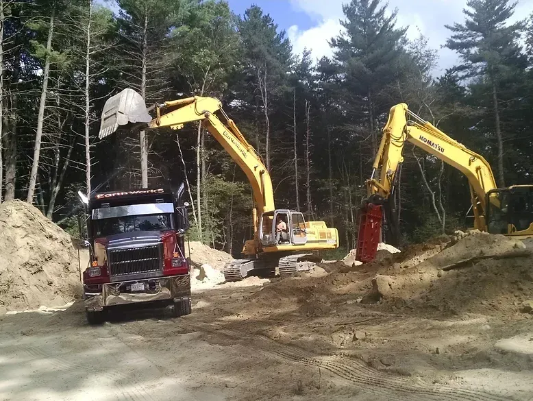Two yellow excavators work in a dirt-filled construction site; one loads a dump truck, while the other digs in the dirt.