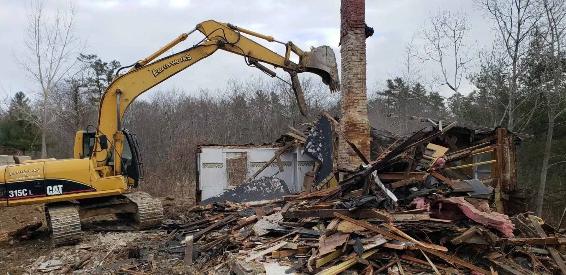 A yellow CAT excavator demoishes a building, with its bucket poised near a tall stone chimney amidst piles of debris.