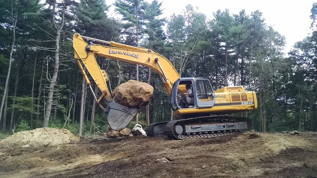 A yellow excavator lifting a large rock at a wooded construction site.