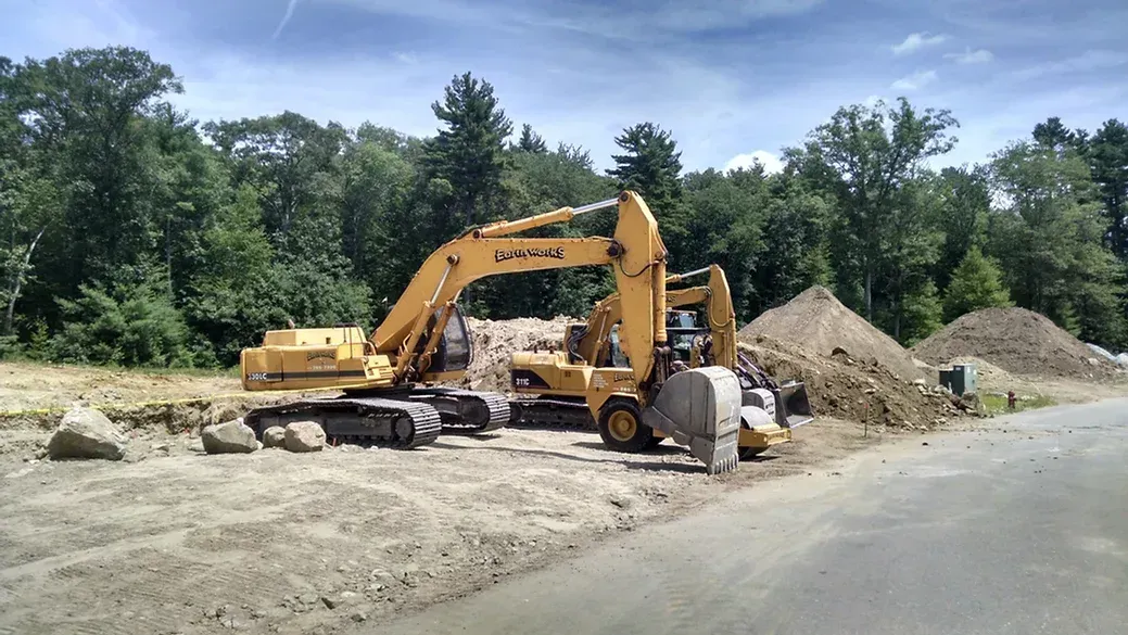 Two yellow excavators work on a dirt construction site with a forest in the background under a blue sky.