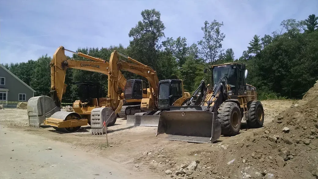 Yellow excavators and a front-end loader parked on a construction site with a house and trees in the background.