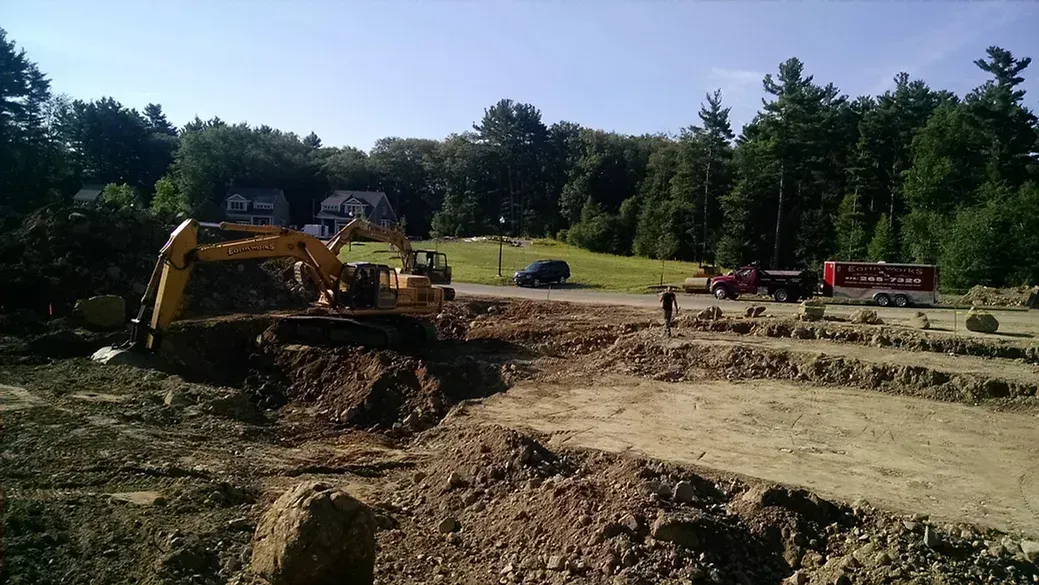 A yellow excavator works on a construction site with piles of dirt, a red container, and trees in the background.