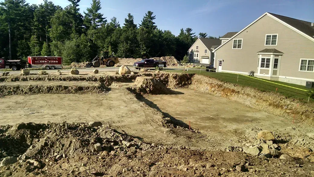Excavated dirt lot prepared for a building foundation next to a finished suburban home and forest.