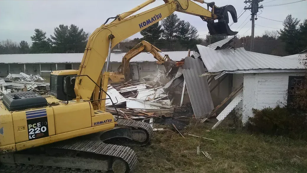 A yellow Komatsu excavator demolishes the roof of a small building with a metal claw.